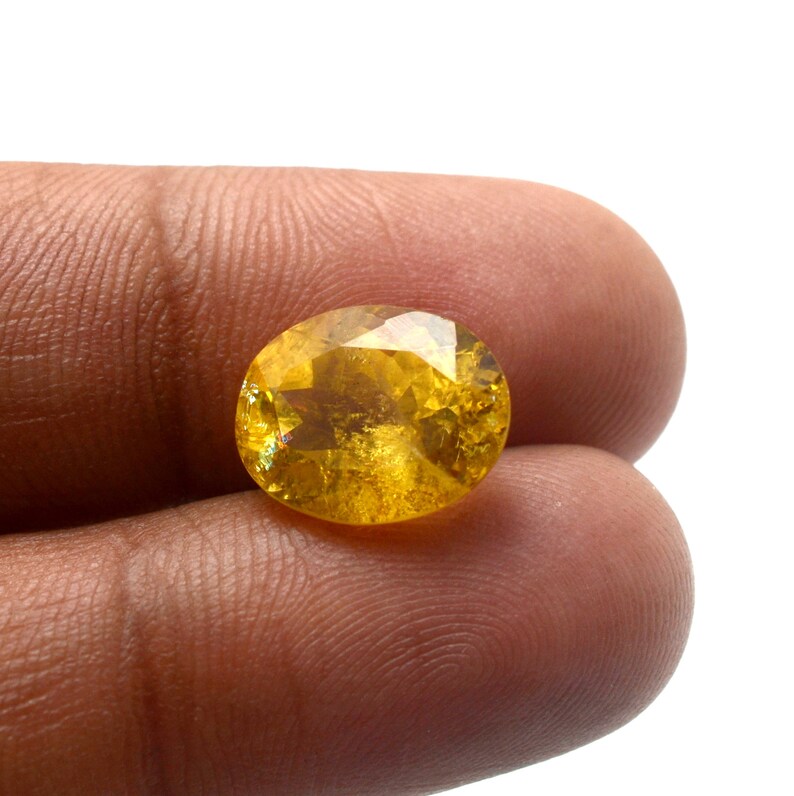 May include: A faceted, oval-shaped gemstone with a vibrant yellow hue. The gem is held between fingers, showcasing its polished surface and internal clarity. The gemstone's facets catch the light, creating a sparkling effect. The gem is likely a citrine or a similar type of gemstone.