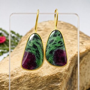 May include: A pair of drop earrings featuring teardrop-shaped zoisite stones with vibrant green and deep magenta inclusions. Each stone is encased in a thin gold-toned metal bezel and attached to a matching gold-toned curved ear wire hook.