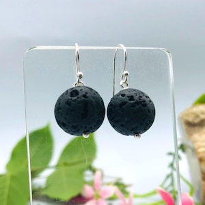 May include: A pair of drop earrings featuring round, porous black lava stone beads suspended from silver-toned metal hooks. The beads have a textured, cratered surface, and the silver hooks are curved for insertion into the earlobe.