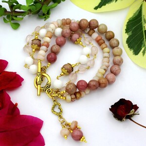May include: A multi-strand beaded bracelet featuring round stones in shades of pink, tan, and white. The bracelet includes gold-toned metal accents, a toggle clasp, and a small heart-shaped charm dangling from the closure mechanism.
