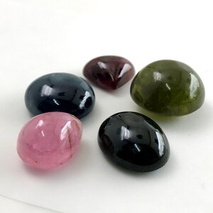 May include: A collection of six polished gemstones in various colors. The stones include a deep blue, a dark red, a green, a black, and a pink stone. The gemstones are oval-shaped and have a glossy finish.