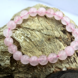 Peut inclure: Un bracelet de perles en quartz rose. Le bracelet est composé de perles rondes rose clair enfilées sur un cordon rose. Les perles ont un aspect légèrement translucide. Le bracelet est présenté sur un morceau de bois vieilli.