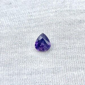 May include: A faceted, pear-shaped, deep purple gemstone. The gem has a brilliant cut, reflecting light with multiple facets. The stone is set against a light gray, textured fabric background. The gemstone is a rich, saturated color.