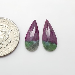 May include: Two pear-shaped, faceted gemstones featuring a dual-tone color pattern of deep magenta-pink and mottled green with dark speckles. A United States quarter coin is positioned to the left for scale, showing the relative size of the stones.