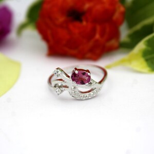 Könnte beinhalten: Ein silberner Ring mit einem rosa Edelstein in der Mitte und kleinen, klaren Steinen. Der Ring hat ein einzigartiges, fließendes Design. Der Hintergrund umfasst eine rote Blume, grüne Blätter und ein gelbes Blütenblatt.