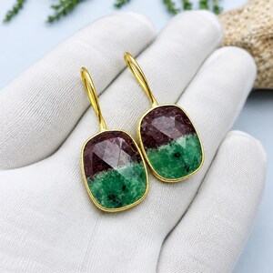 Pode incluir: Um par de brincos pendentes com pedras retangulares facetadas de cantos arredondados. Cada pedra apresenta uma divisão de cor distinta entre vermelho profundo e verde vibrante, engastadas numa armação de metal dourado com gancho.