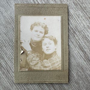 Puede incluir: Fotografía en tonos sepia de dos mujeres en un marco decorativo. Una mujer lleva un top oscuro y la otra una camisa a cuadros. El marco tiene un borde texturizado y la foto muestra signos de envejecimiento.