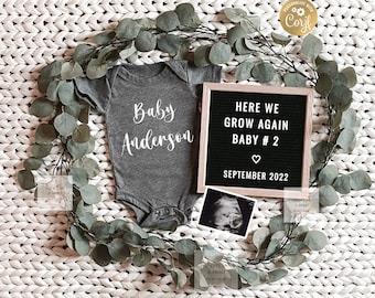 May include: A gray onesie with the text "Baby Anderson" on it, a black letter board with the text "Here we grow again Baby #2 September 2022", and a small ultrasound image. The items are arranged on a white knitted surface with a green garland around them.