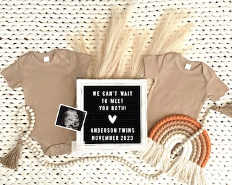 May include: Two tan baby onesies, a letter board with the text "We can't wait to meet you both! Anderson Twins November 2023", a small ultrasound image, a wooden beaded garland with a tassel, and a woven rainbow with tassels.