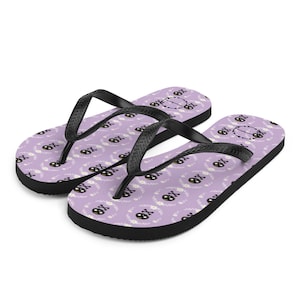 May include: A pair of black flip-flops with a purple background and a repeating pattern of black skulls with white flowers. The skulls have a white circle around them with the letters "OX" inside.
