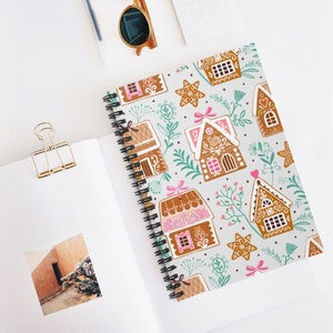 May include: A spiral bound notebook with a white background and a repeating pattern of gingerbread houses, stars, and floral designs. The gingerbread houses are decorated with pink and white icing and candy.