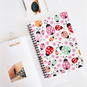 Puede incluir: Un cuaderno con espiral con un patrón de mariquitas de colores. La portada presenta mariquitas rojas, rosas, naranjas y verdes, junto con flores y corazones rosas sobre un fondo blanco. El cuaderno está abierto, revelando páginas en blanco.