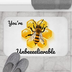 May include: White bath mat with a bee on a honeycomb design. The text "You're Unbeelievable" is written in black cursive.