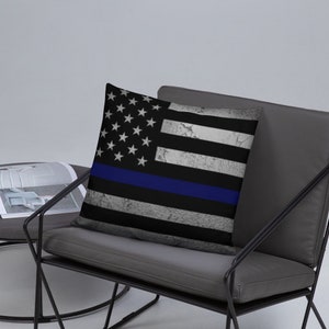 Puede incluir: Una almohada decorativa con un diseño de bandera estadounidense desgastada con una fina línea azul. La almohada está sobre una silla gris con patas de metal negro.