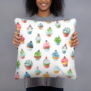 May include: A white throw pillow with a colorful watercolor pattern of cupcakes. The cupcakes are decorated with different colors of frosting and sprinkles.