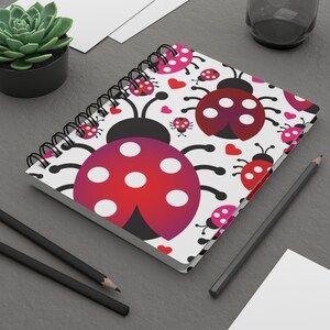 Puede incluir: Una libreta de espiral blanca con un patrón repetido de mariquitas rojas y rosas con puntos blancos y corazones rosas. Las mariquitas tienen patas y antenas negras.