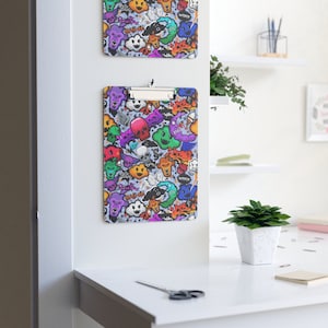 May include: A colorful clipboard with a graphic design of cartoon skulls, clouds, and other whimsical characters. The clipboard is made of plastic and has a metal clip.