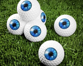 May include: Six white golf balls with blue eyes printed on them. The golf balls are arranged in a pyramid shape on a green grassy surface.