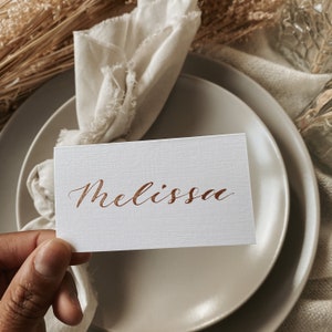 May include: A white place card with the name "Melissa" written in copper calligraphy.