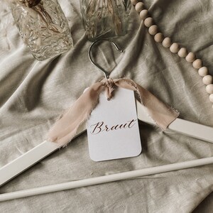 May include: A white wooden hanger with a pink ribbon and a white card that says "Braut" in brown calligraphy.