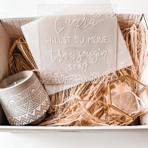 May include: A white box filled with straw, a small gray candle with a geometric pattern, a clear acrylic sign with the text "Laura, willst du meine Trauzeugin sein?" and a small gold box with a pink tulle bag inside.