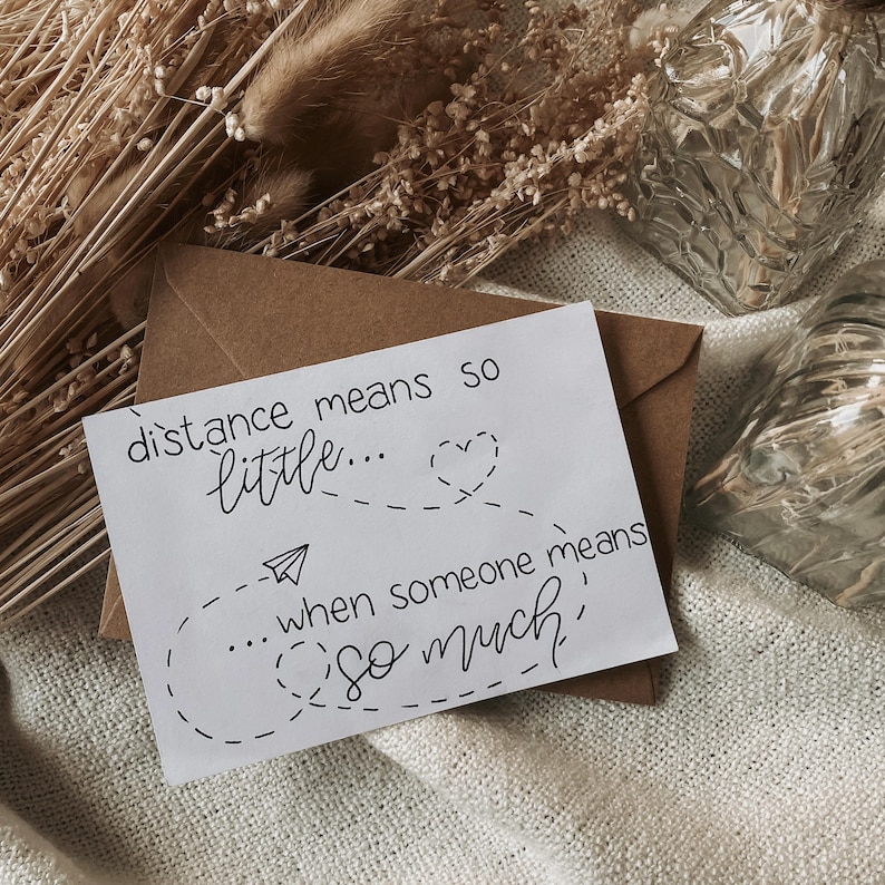 K&ouml;nnte beinhalten: Eine wei&szlig;e Karte mit dem handgeschriebenen Text "distance means so little... when someone means so much" in schwarzer Tinte. Die Karte liegt auf einem braunen Umschlag, mit getrockneten Blumen im Hintergrund.
