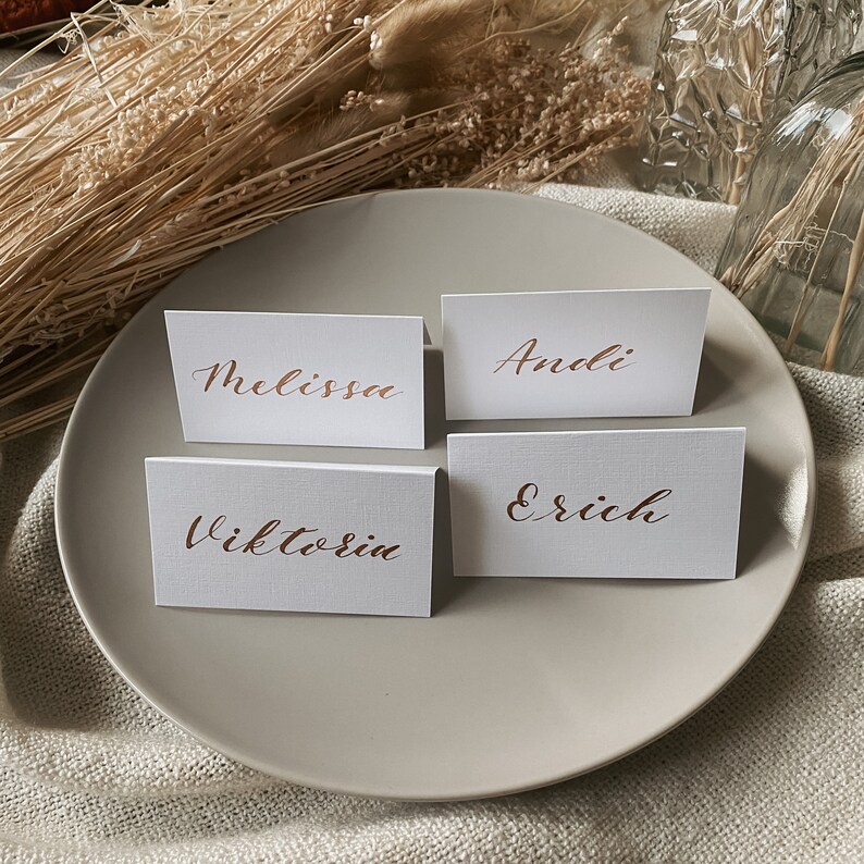 May include: Four white place cards with copper calligraphy on a white plate. The names on the cards are Melissa, Andi, Viktorin, and Erich.