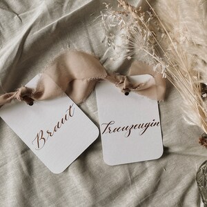May include: Two white cards with brown ribbon tied around them. The cards have the words "Braut" and "Trauzeugin" written in calligraphy on them.