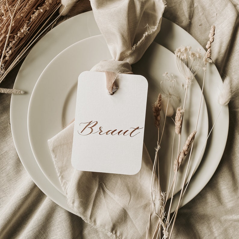 May include: A white card with the word "Braut" written in calligraphy on it. The card is placed on a beige linen napkin and is surrounded by dried flowers and a white plate.