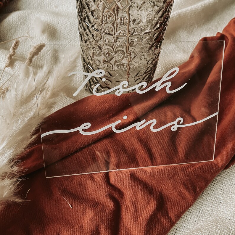 May include: A clear acrylic sign with the text "Tisch eins" written in white calligraphy. The sign is sitting on a red fabric surface.