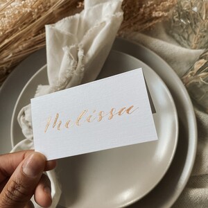 May include: A white place card with the name "Melissa" written in gold calligraphy.