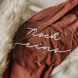May include: A clear acrylic sign with the text "Tisch eins" written in white cursive script. The sign is sitting on a brown fabric surface.