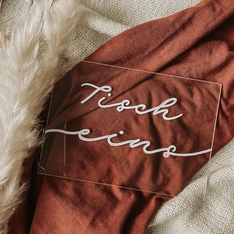 May include: A clear acrylic sign with the text "Tisch eins" written in white cursive script. The sign is sitting on a brown fabric surface.