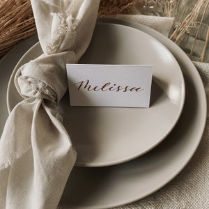 May include: A place setting with a white place card with the name "Melissa" written in brown calligraphy. The place card is on top of a beige plate. A beige napkin is tied around the plate.
