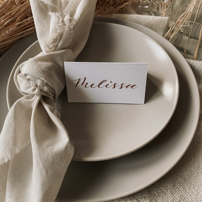 May include: A place setting with a white place card with the name "Melissa" written in brown calligraphy. The place card is on top of a beige plate. A beige napkin is tied around the plate.