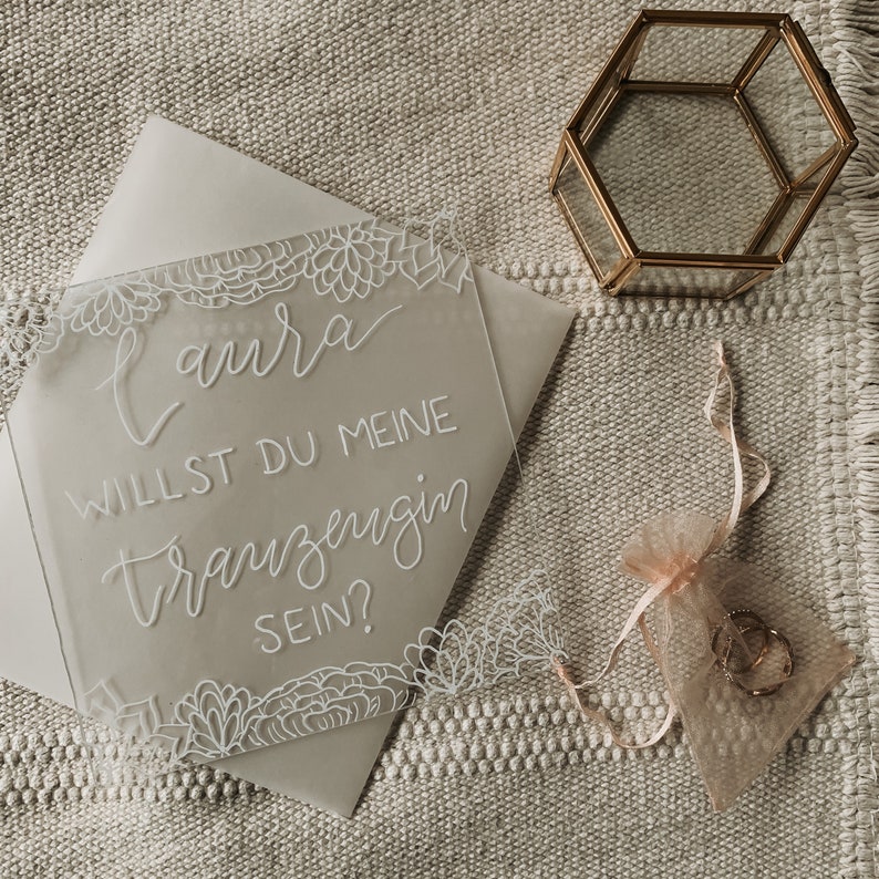 May include: A white acrylic sign with a floral design and the text "Laura, willst du meine Trauzeugin sein?"  The sign is on a beige textured surface.