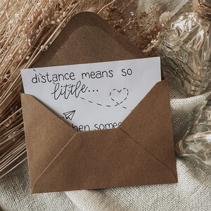 K&ouml;nnte beinhalten: Ein brauner Papierumschlag, der eine wei&szlig;e Karte enth&auml;lt. Auf der Karte steht in schwarzer Schrift "distance means so little...", erg&auml;nzt durch eine gestrichelte Linie zu einem Herz und einer Papierflugzeug-Illustration. Der Umschlag ist ge&ouml;ffnet.