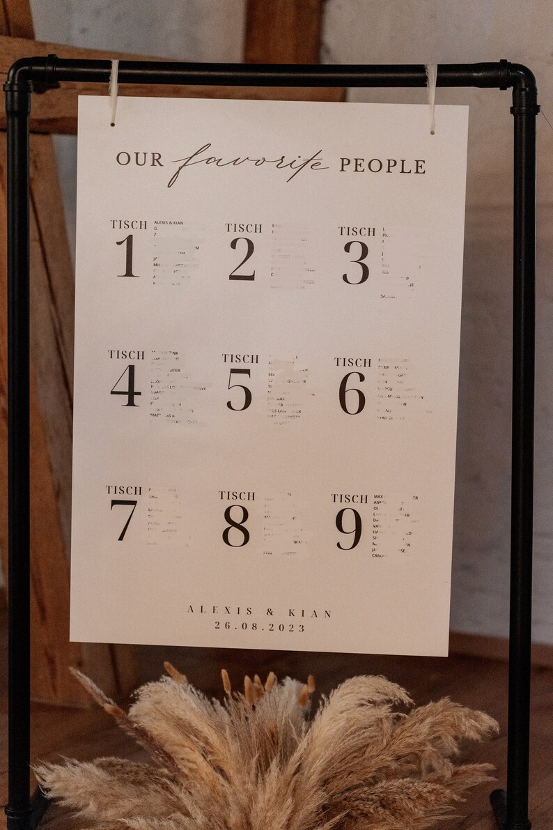 May include: A white sign with black text that reads "OUR favorite PEOPLE" and lists table numbers 1 through 9. The sign is hanging on a black metal stand. The text at the bottom of the sign reads "ALEXIS & KIAN 26.08.2023".