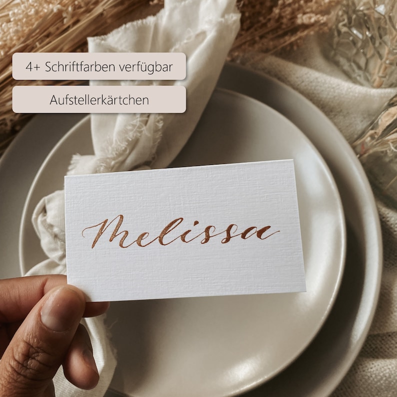 May include: A white place card with the name "Melissa" written in copper calligraphy. The card is on a beige plate with a white napkin folded on top. The text "4+ Schriftfarben verf&uuml;gbar" and "Aufstellerk&auml;rtchen" is visible in the background.