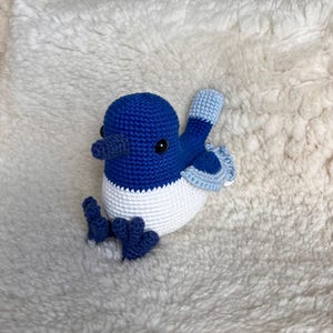 May include: A handmade crochet bird in shades of blue and white. The bird has a dark blue head and feet, a white body, and light blue wings. The bird is sitting on a fluffy, white surface.