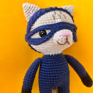May include: A handmade crochet cat doll in a superhero costume. The doll has a white face, a blue mask, and a blue jumpsuit with black gloves and boots. The cat has black eyes and a pink nose. The background is a solid yellow.