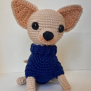 May include: A crocheted Chihuahua dog figurine with large ears and a dark blue sweater. The dog has black eyes and a black nose. The ears and paws are a light tan color. The figurine is handmade and sits upright.