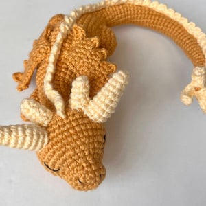 Könnte beinhalten: Ein gehäkeltes Drachenspielzeug mit einem hellbraunen Körper und einem cremefarbenen Kopf und Schwanz. Der Drache hat einen geschlossenen Mund und einen eingerollten Schwanz.