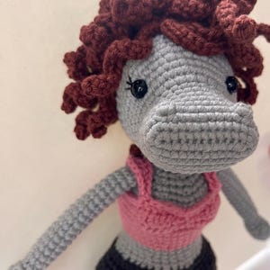 May include: Crocheted hippo doll with gray body, dark red curly hair, and black eyes. The hippo wears a pink top and black pants. The doll's arms are outstretched, and it has a friendly expression. The handmade toy is a unique gift.