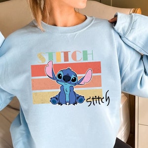 May include: Light blue sweatshirt with a retro-style graphic of Stitch from Lilo & Stitch. The graphic features Stitch sitting with his arms raised and the text "Stitch" in a colorful, retro font.
