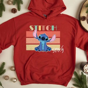 May include: Red hooded sweatshirt with a retro-style graphic of Stitch from Lilo & Stitch. The graphic features Stitch with a blue and light blue color scheme and the text "Stitch" and "Stitchy" in a rainbow color scheme.