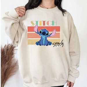 May include: A beige sweatshirt with a retro-style graphic of Stitch from Lilo & Stitch. The graphic features Stitch sitting with his arms crossed, surrounded by colorful stripes. The text "Stitch" is printed above and below the graphic.