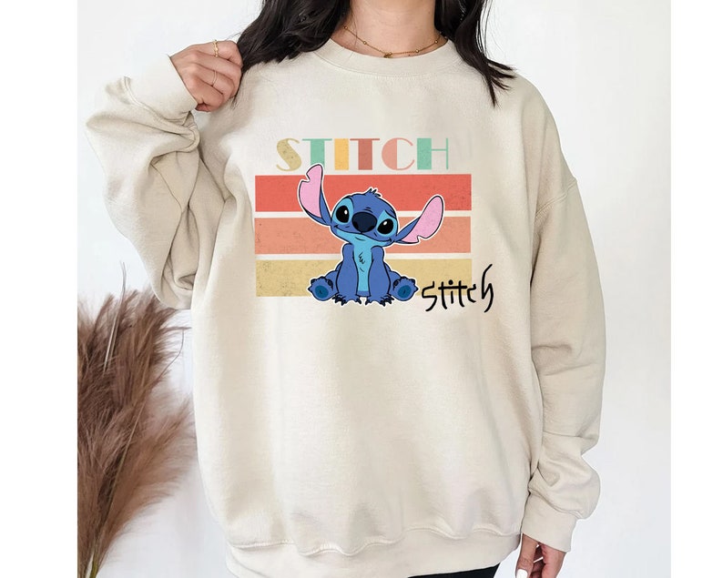 May include: A beige sweatshirt with a retro-style graphic of Stitch from Lilo & Stitch. The graphic features Stitch sitting with his arms crossed, surrounded by colorful stripes. The text "Stitch" is printed above and below the graphic.