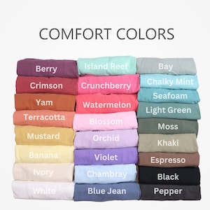 May include: A stack of folded t-shirts in various colors, including berry, crimson, yam, terracotta, mustard, banana, ivory, white, island reef, crunchberry, watermelon, blossom, orchid, violet, chambray, blue jean, bay, chalky mint, seafoam, light green, moss, khaki, espresso, black, and pepper. The text "COMFORT COLORS" is at the top of the image.