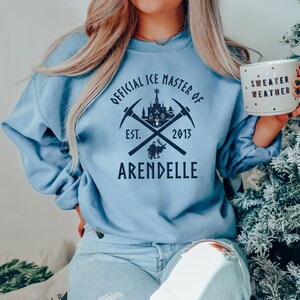 May include: A light blue sweatshirt with a graphic design featuring a castle, crossed pickaxes, and the text "Official Ice Master of Arendelle Est. 2013".